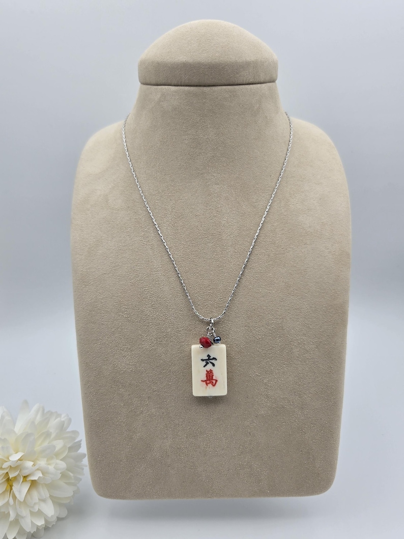 May include: A silver-toned chain necklace featuring a rectangular off-white mahjong tile pendant. The tile displays the black Chinese character for six and the red character for ten thousand. A small red bead and a tiny blue bead accent the top.
