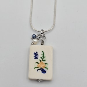 May include: A silver snake chain necklace featuring a rectangular off-white mahjong tile pendant. The tile displays a blue Japanese character and a painted yellow and green floral design. Two small beads, one blue and one white, hang from the silver bail.