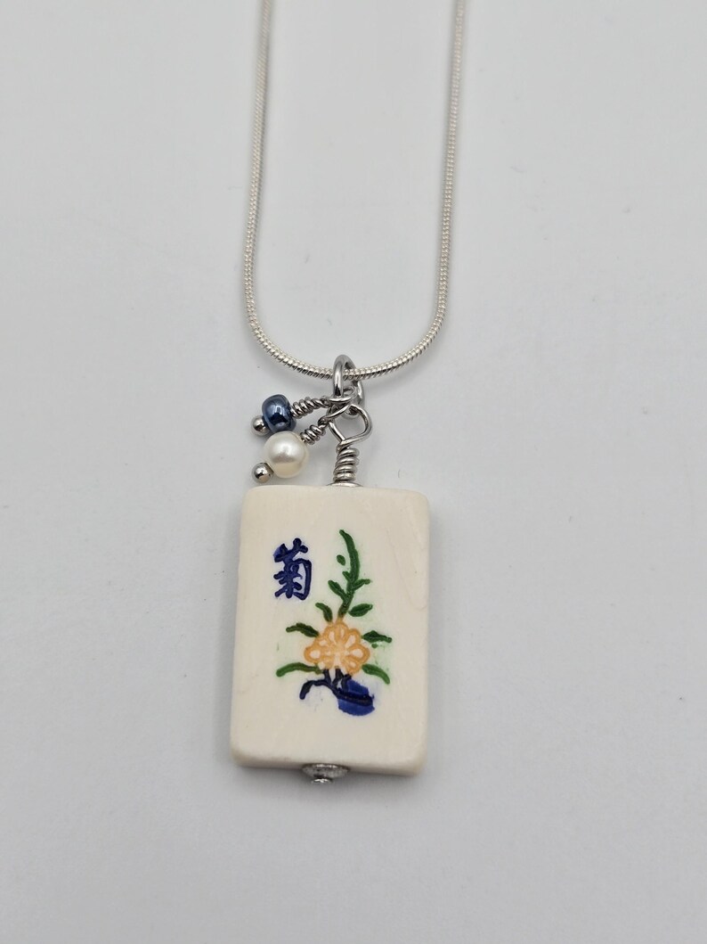 May include: A silver snake chain necklace featuring a rectangular off-white mahjong tile pendant. The tile displays a blue Japanese character and a painted yellow and green floral design. Two small beads, one blue and one white, hang from the silver bail.