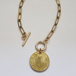 May include: A gold chain bracelet with a gold coin charm featuring the Irish harp and the year 2001. The chain is made of oval links and has a toggle clasp.