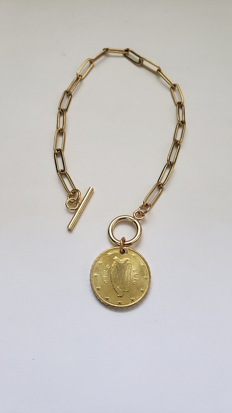 May include: A gold chain bracelet with a gold coin charm featuring the Irish harp and the year 2001. The chain is made of oval links and has a toggle clasp.