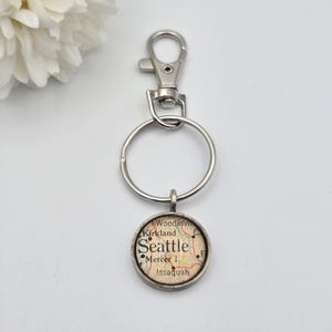 May include: A silver-toned keychain with a round pendant featuring a map of Seattle, Washington. The pendant includes the city name and surrounding areas like Mercer Island and Issaquah. The keychain has a clasp and a split ring.