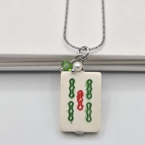 May include: A silver-toned chain necklace featuring a rectangular off-white mahjong tile pendant. The tile is decorated with green and red engraved symbols. A small green crystal bead and a white pearl bead are attached to the silver jump ring.