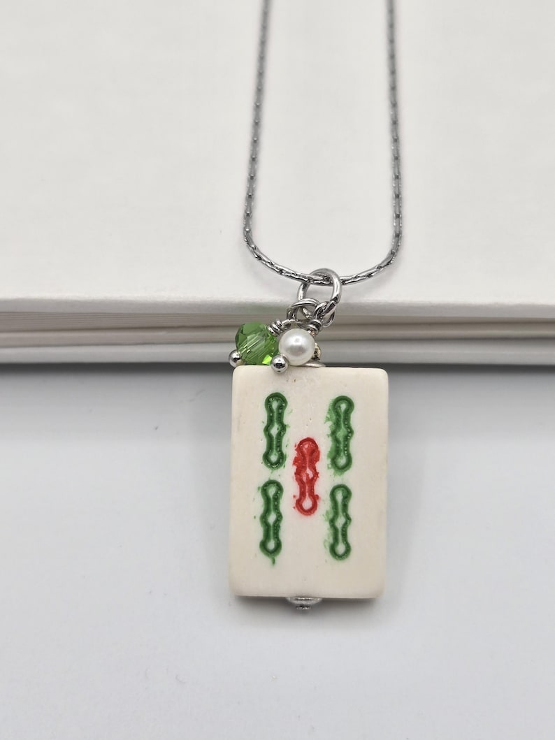 May include: A silver-toned chain necklace featuring a rectangular off-white mahjong tile pendant. The tile is decorated with green and red engraved symbols. A small green crystal bead and a white pearl bead are attached to the silver jump ring.