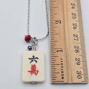 May include: A silver-toned chain necklace featuring a rectangular off-white mahjong tile pendant with black and red Chinese characters. The pendant is accented with a small red faceted bead and a dark blue bead. A wooden ruler shows the tile is 2.5 cm long.