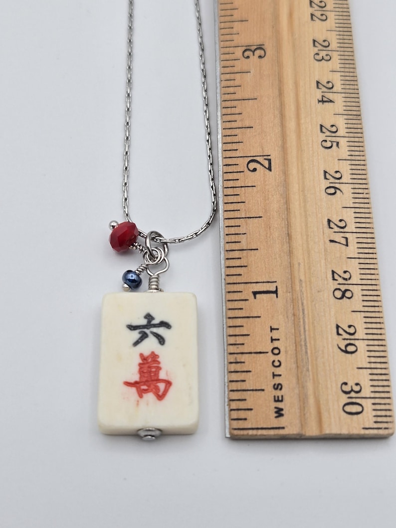May include: A silver-toned chain necklace featuring a rectangular off-white mahjong tile pendant with black and red Chinese characters. The pendant is accented with a small red faceted bead and a dark blue bead. A wooden ruler shows the tile is 2.5 cm long.