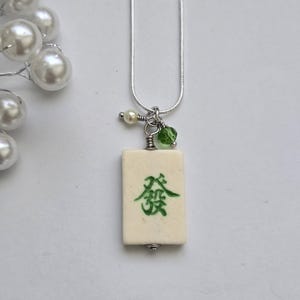 May include: A silver-toned snake chain necklace featuring a rectangular off-white mahjong tile pendant with a green Chinese character. The pendant is accented with a small white pearl bead and a translucent green crystal bead attached to the silver bail.