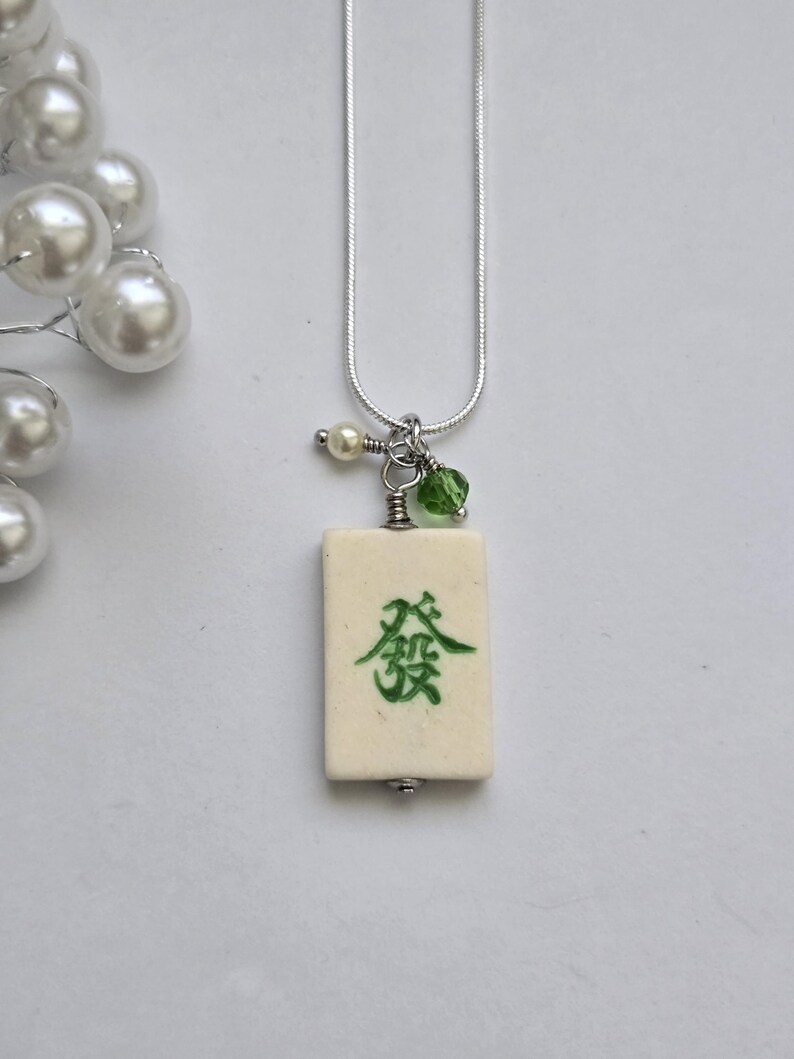 May include: A silver-toned snake chain necklace featuring a rectangular off-white mahjong tile pendant with a green Chinese character. The pendant is accented with a small white pearl bead and a translucent green crystal bead attached to the silver bail.