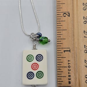 May include: A silver-tone snake chain necklace featuring a rectangular off-white mahjong tile pendant with five circular symbols in green, blue, and red. A small green faceted bead and a dark blue bead hang above the tile next to a wooden ruler.