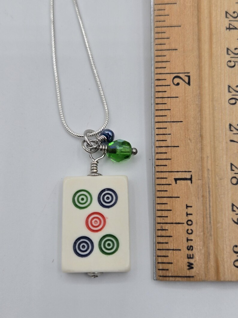 May include: A silver-tone snake chain necklace featuring a rectangular off-white mahjong tile pendant with five circular symbols in green, blue, and red. A small green faceted bead and a dark blue bead hang above the tile next to a wooden ruler.