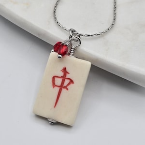 May include: A silver-toned chain necklace featuring a rectangular off-white mahjong tile pendant. The tile is engraved with a red Chinese character and is accented by two small, faceted red beads attached to the silver wire bail at the top.
