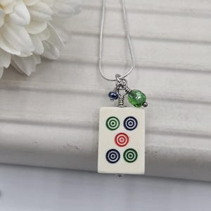 May include: A silver-toned chain necklace featuring a rectangular off-white mahjong tile pendant. The tile displays five circular symbols in green, blue, and red. Two small accent beads, one dark blue and one faceted lime green, hang above the tile.