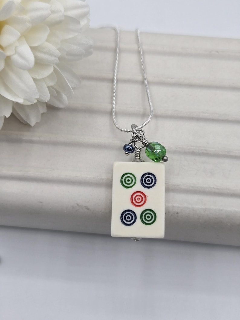 May include: A silver-toned chain necklace featuring a rectangular off-white mahjong tile pendant. The tile displays five circular symbols in green, blue, and red. Two small accent beads, one dark blue and one faceted lime green, hang above the tile.