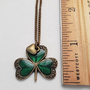 May include: A gold chain necklace with a green shamrock pendant. The shamrock has a small gold heart charm attached to it. The pendant is approximately 2.5 cm in diameter.