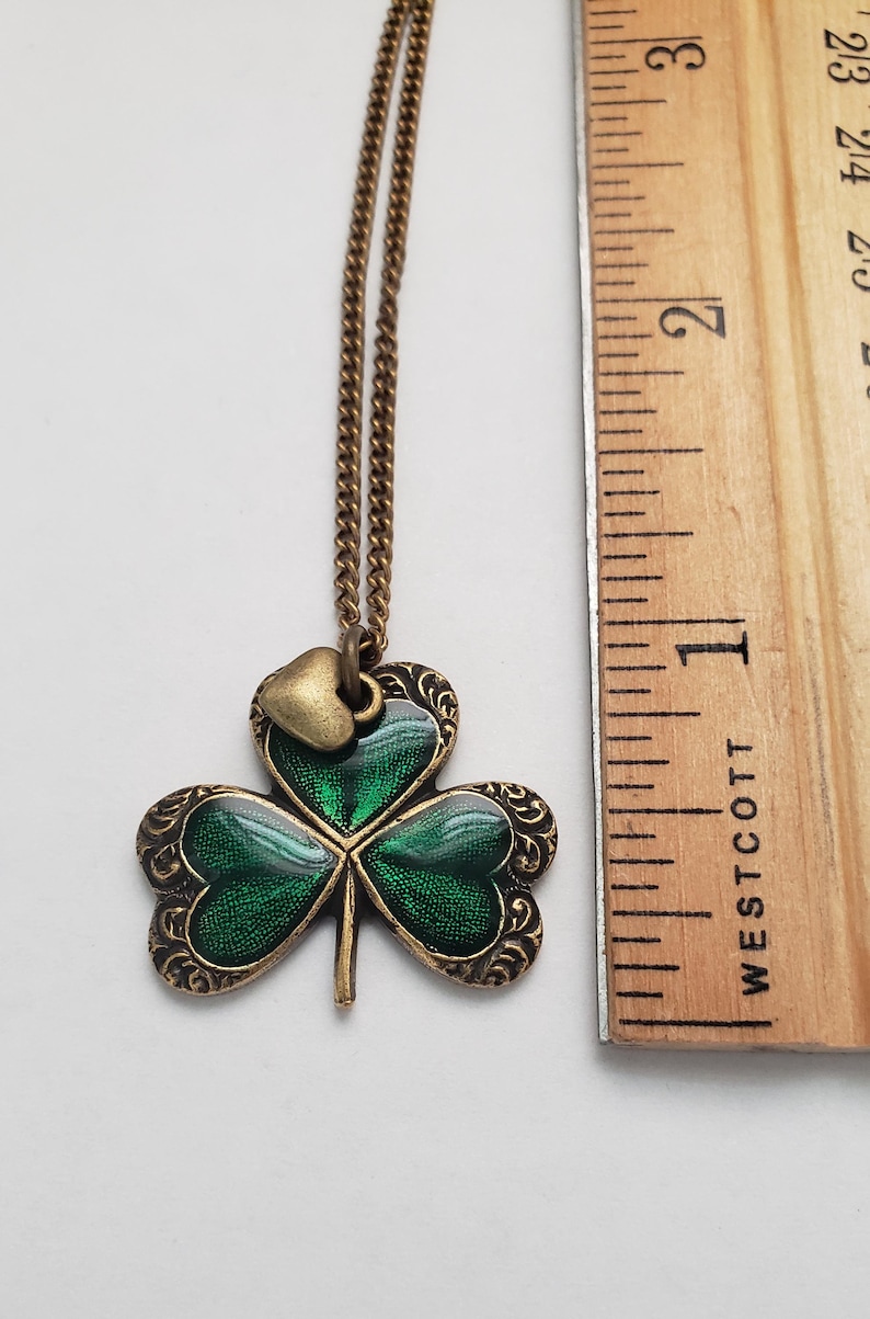 May include: A gold chain necklace with a green shamrock pendant. The shamrock has a small gold heart charm attached to it. The pendant is approximately 2.5 cm in diameter.