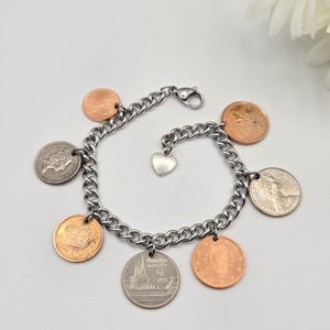 May include: A silver-toned metal chain link bracelet featuring multiple dangling coins of varying sizes and colors, including copper and silver tones. A small, plain silver-toned heart charm is attached to the center of the chain, which secures with a lobster claw clasp.