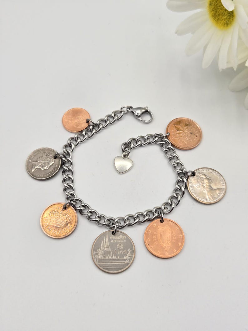 May include: A silver-toned metal chain link bracelet featuring multiple dangling coins of varying sizes and colors, including copper and silver tones. A small, plain silver-toned heart charm is attached to the center of the chain, which secures with a lobster claw clasp.