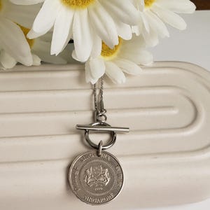 May include: Silver necklace with a Singapore dollar coin pendant and a toggle clasp. The coin features the Singapore coat of arms and the year 1990.