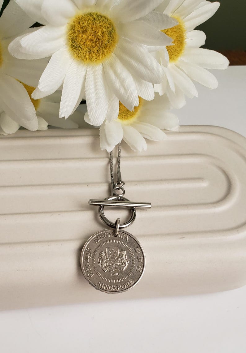 May include: Silver necklace with a Singapore dollar coin pendant and a toggle clasp. The coin features the Singapore coat of arms and the year 1990.