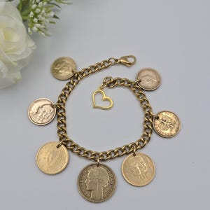 May include: A gold-toned chain link charm bracelet featuring six circular coin charms and one open heart-shaped charm. The coins display various profiles and symbols, including the text REPUBLIQUE FRANCAISE and 20F. It has a lobster claw clasp.