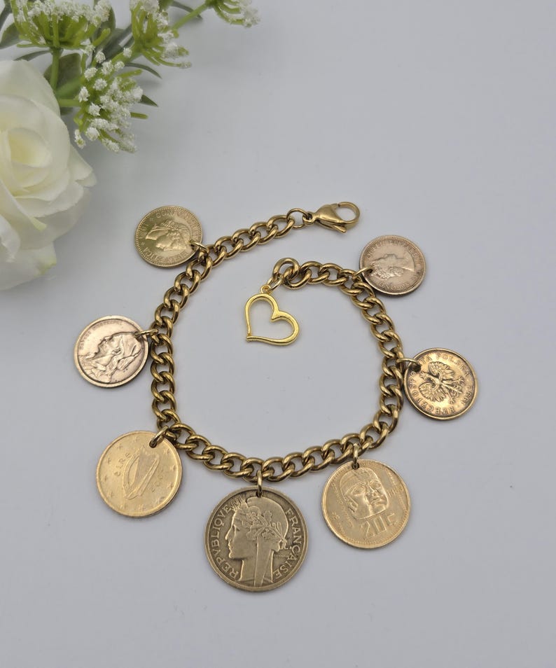 May include: A gold-toned chain link charm bracelet featuring six circular coin charms and one open heart-shaped charm. The coins display various profiles and symbols, including the text REPUBLIQUE FRANCAISE and 20F. It has a lobster claw clasp.