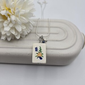 May include: A rectangular off-white mahjong tile pendant featuring a painted yellow flower and blue Chinese character, suspended from a silver-toned chain. The pendant includes a small wire-wrapped accent with a tiny blue bead and a white pearl.