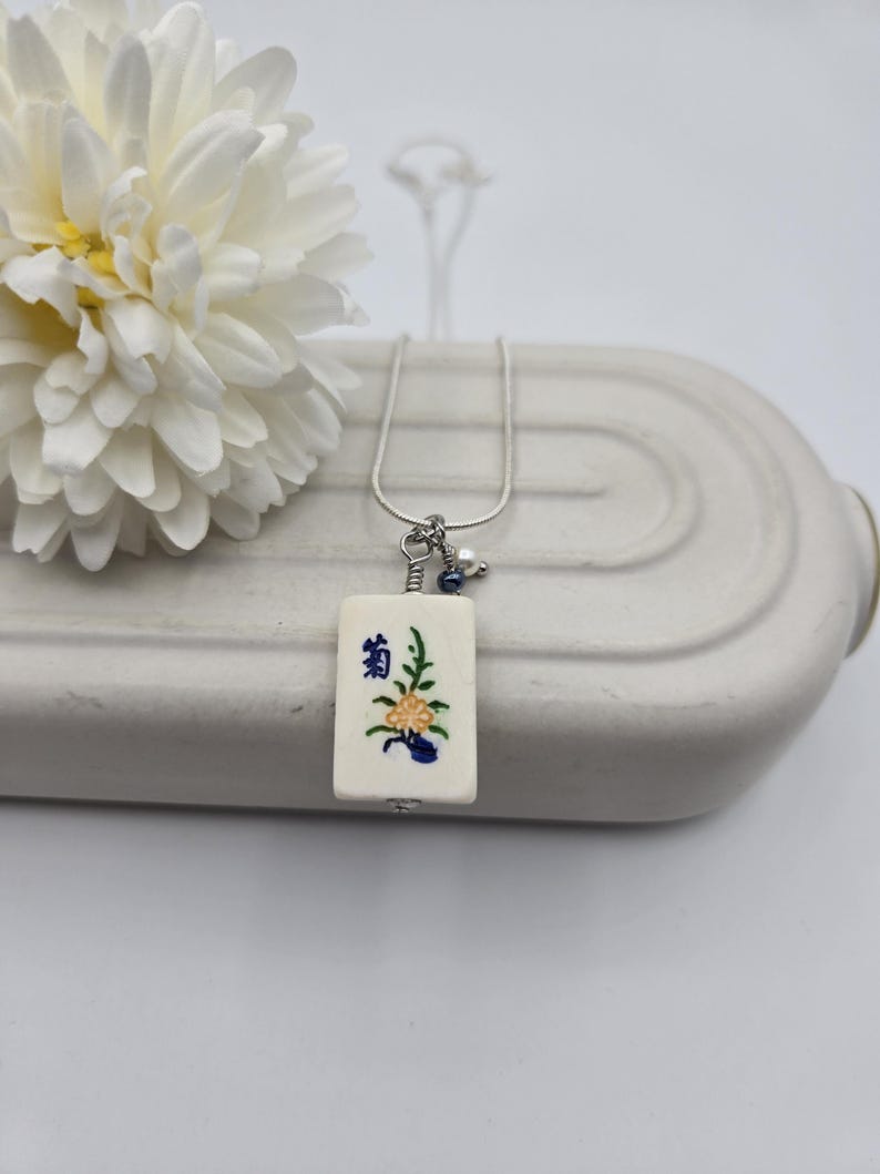 May include: A rectangular off-white mahjong tile pendant featuring a painted yellow flower and blue Chinese character, suspended from a silver-toned chain. The pendant includes a small wire-wrapped accent with a tiny blue bead and a white pearl.