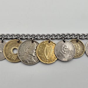 Pode incluir: Uma pulseira de corrente de metal prateado com uma s&eacute;rie de moedas internacionais pendentes. As moedas variam de cor entre tons dourados e prateados, exibindo diversos retratos, uma harpa e caracteres japoneses, presas por pequenos an&eacute;is.
