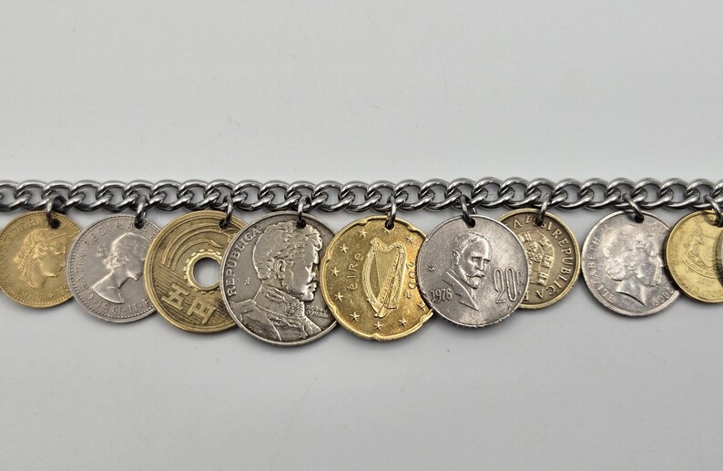 Pode incluir: Uma pulseira de corrente de metal prateado com uma s&eacute;rie de moedas internacionais pendentes. As moedas variam de cor entre tons dourados e prateados, exibindo diversos retratos, uma harpa e caracteres japoneses, presas por pequenos an&eacute;is.