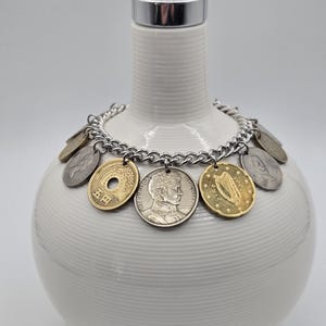 Pode incluir: Uma pulseira de corrente de metal prateado com v&aacute;rias moedas internacionais como pingentes, incluindo uma moeda japonesa de cinco ienes, uma moeda chilena com um retrato e uma moeda irlandesa com um design de harpa, dispostas em c&iacute;rculo.