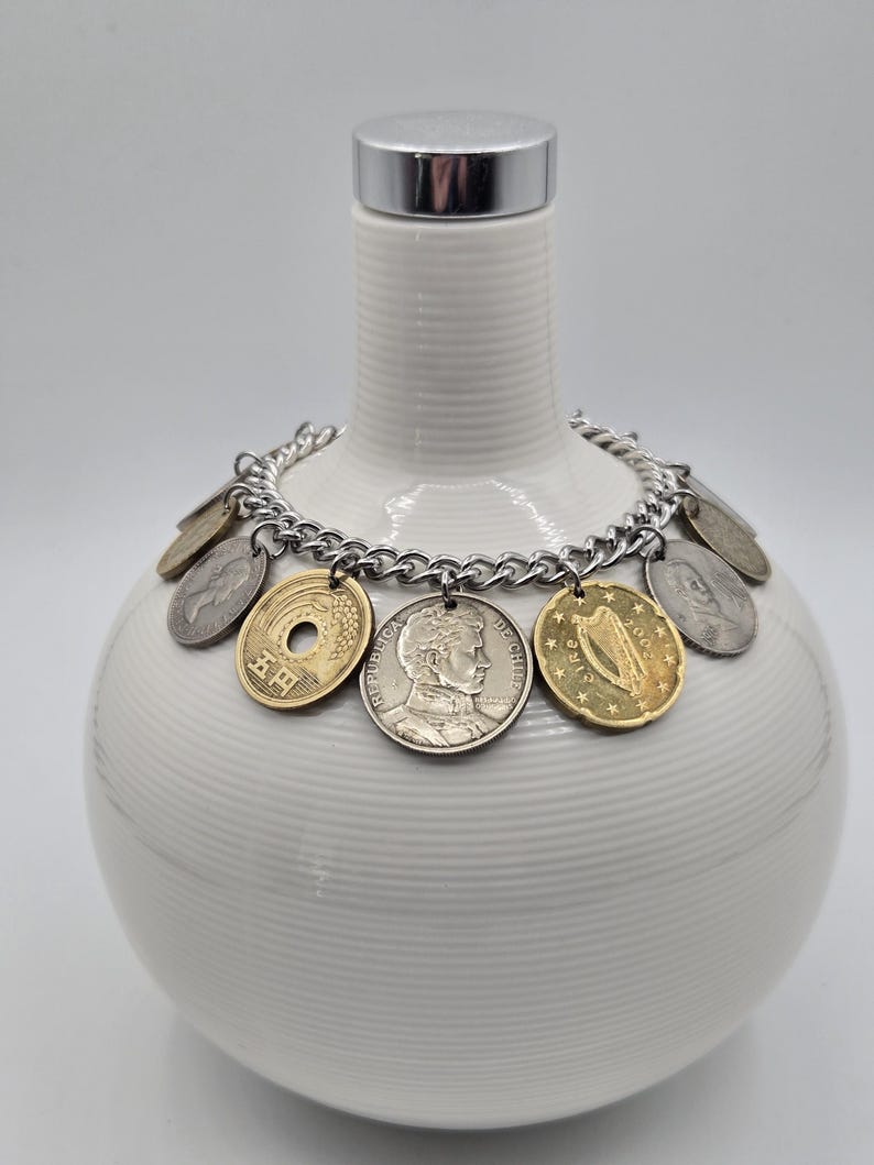 Pode incluir: Uma pulseira de corrente de metal prateado com v&aacute;rias moedas internacionais como pingentes, incluindo uma moeda japonesa de cinco ienes, uma moeda chilena com um retrato e uma moeda irlandesa com um design de harpa, dispostas em c&iacute;rculo.