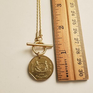 May include: A gold necklace with a Singaporean coin pendant. The coin features the Singapore coat of arms and the year 1995. The necklace has a toggle clasp.