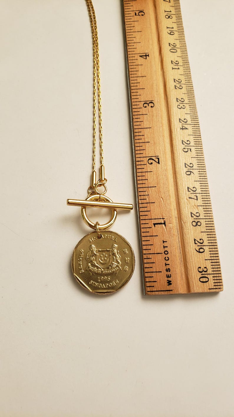 May include: A gold necklace with a Singaporean coin pendant. The coin features the Singapore coat of arms and the year 1995. The necklace has a toggle clasp.