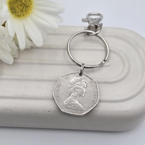 May include: A silver coin keychain featuring a coin with a profile of a person. The keychain has a silver ring and clasp. A white flower with a yellow center is in the background.