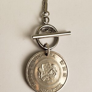 May include: A silver necklace with a toggle clasp and a Singapore dollar coin pendant. The coin features the Singapore coat of arms and the year 1990.