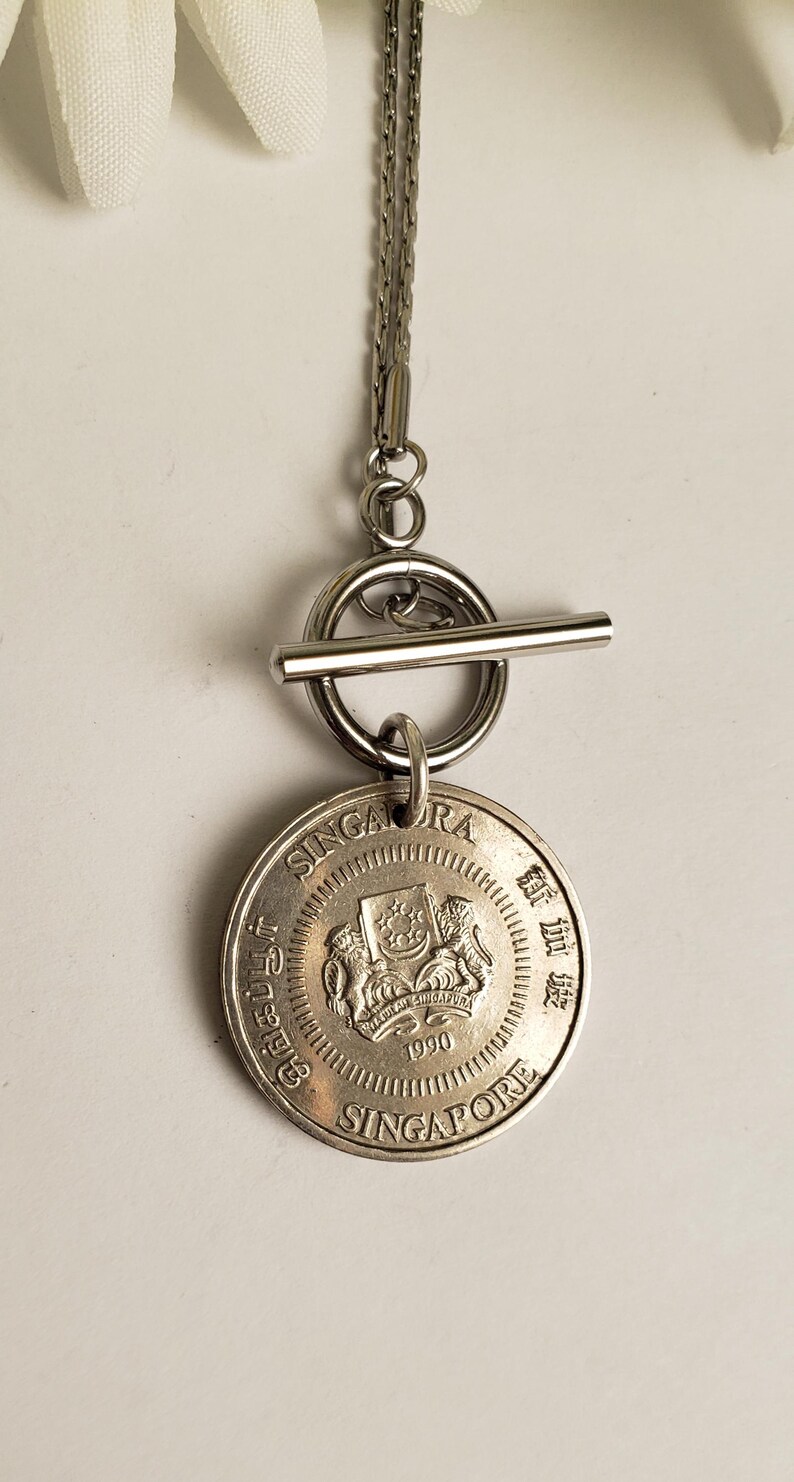 May include: A silver necklace with a toggle clasp and a Singapore dollar coin pendant. The coin features the Singapore coat of arms and the year 1990.