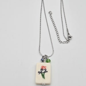 May include: A silver-toned chain necklace featuring a rectangular off-white pendant with a hand-painted red and green bird illustration. The pendant is accented with one small blue bead and one small green bead, attached by a silver jump ring.