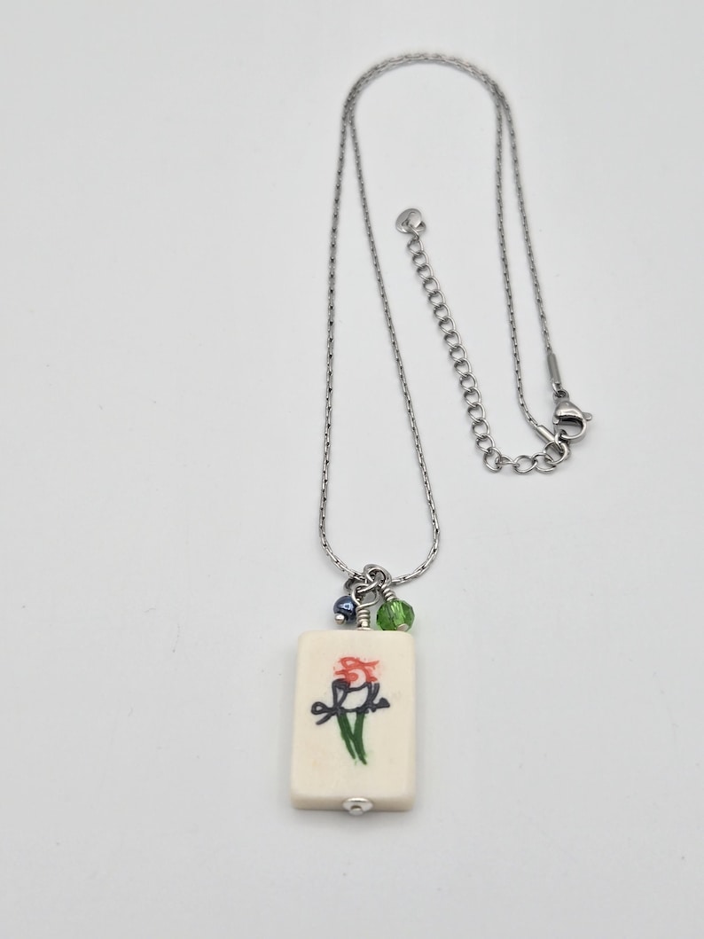May include: A silver-toned chain necklace featuring a rectangular off-white pendant with a hand-painted red and green bird illustration. The pendant is accented with one small blue bead and one small green bead, attached by a silver jump ring.