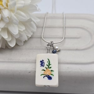 May include: A silver-toned chain necklace featuring a rectangular off-white pendant with a hand-painted yellow flower, green leaves, and blue Japanese kanji characters. The pendant is accented with a small white pearl and a dark blue bead.