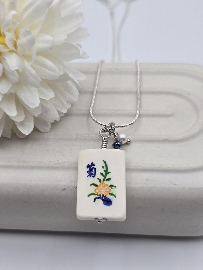 May include: A silver-toned chain necklace featuring a rectangular off-white pendant with a hand-painted yellow flower, green leaves, and blue Japanese kanji characters. The pendant is accented with a small white pearl and a dark blue bead.