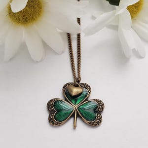 May include: A green shamrock pendant necklace with a small gold heart charm. The necklace is on a gold chain.