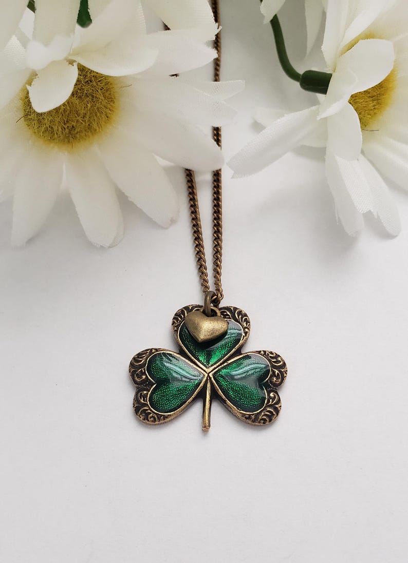May include: A green shamrock pendant necklace with a small gold heart charm. The necklace is on a gold chain.