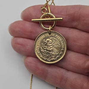 K&ouml;nnte beinhalten: Eine goldfarbene Halskette mit einem runden Anh&auml;nger, der einer mexikanischen M&uuml;nze mit Adleremblem und der Aufschrift ESTADOS UNIDOS MEXICANOS &auml;hnelt. Der Anh&auml;nger ist an einem goldenen Knebelverschluss und einer feinen, strukturierten Kette befestigt.