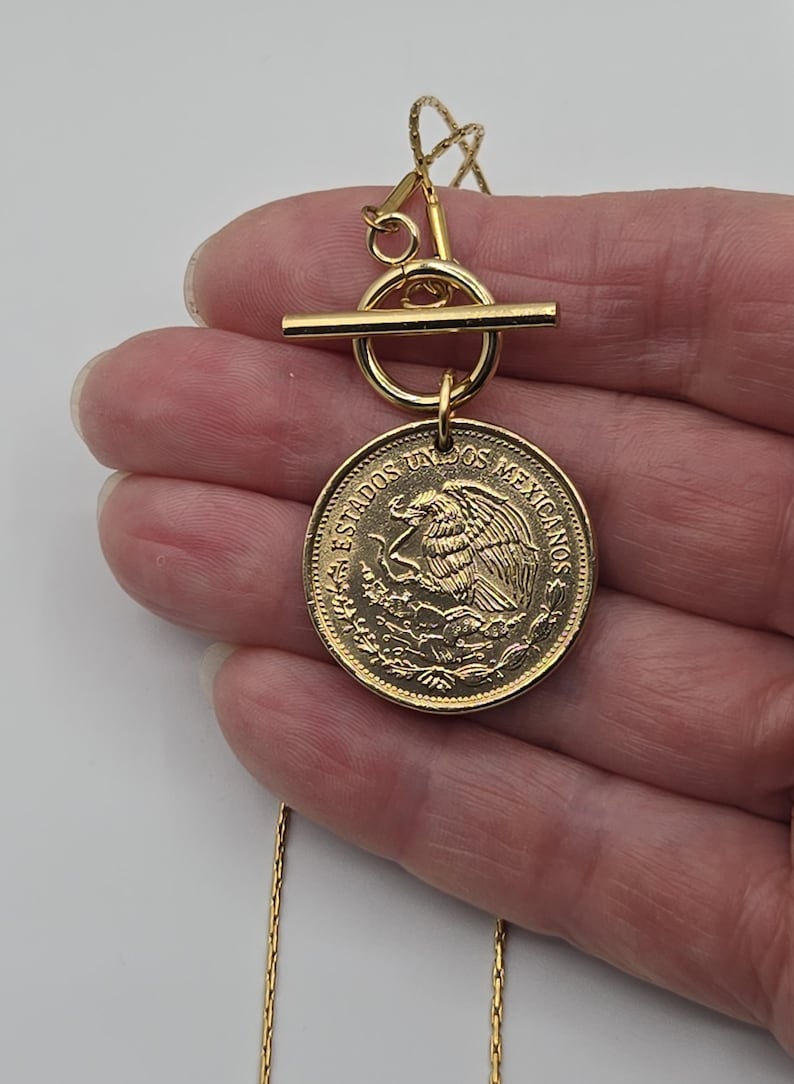 K&ouml;nnte beinhalten: Eine goldfarbene Halskette mit einem runden Anh&auml;nger, der einer mexikanischen M&uuml;nze mit Adleremblem und der Aufschrift ESTADOS UNIDOS MEXICANOS &auml;hnelt. Der Anh&auml;nger ist an einem goldenen Knebelverschluss und einer feinen, strukturierten Kette befestigt.