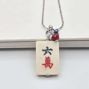 May include: A necklace featuring a rectangular off-white mahjong tile pendant with the black and red Chinese character for six ten-thousands. The pendant is attached to a silver-toned chain with a small red faceted bead and a dark blue bead.