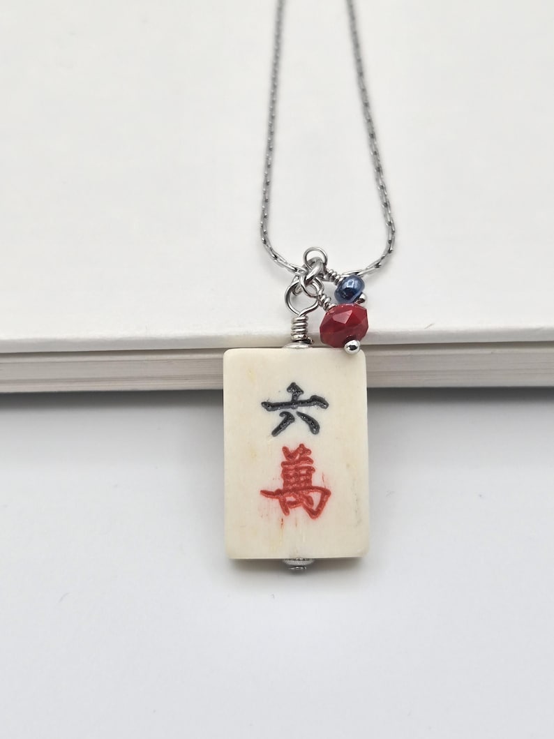 May include: A necklace featuring a rectangular off-white mahjong tile pendant with the black and red Chinese character for six ten-thousands. The pendant is attached to a silver-toned chain with a small red faceted bead and a dark blue bead.