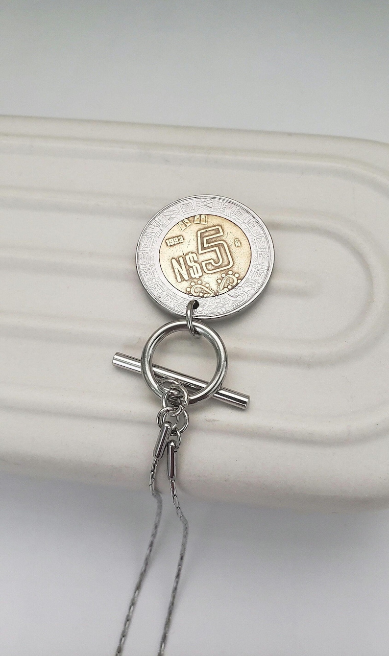 K&ouml;nnte beinhalten: Eine silberfarbene Halskette mit einem runden Knebelverschluss und einem Anh&auml;nger aus einer mexikanischen 5 N$-M&uuml;nze von 1993. Die M&uuml;nze hat eine goldfarbene Mitte und einen silberfarbenen Au&szlig;enring mit gepr&auml;gten Details und dem Text N$5.