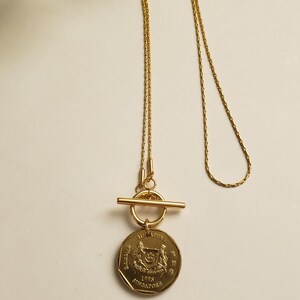 May include: A gold necklace with a toggle clasp and a Singapore dollar coin pendant. The coin features the Singapore coat of arms and the year 1995.
