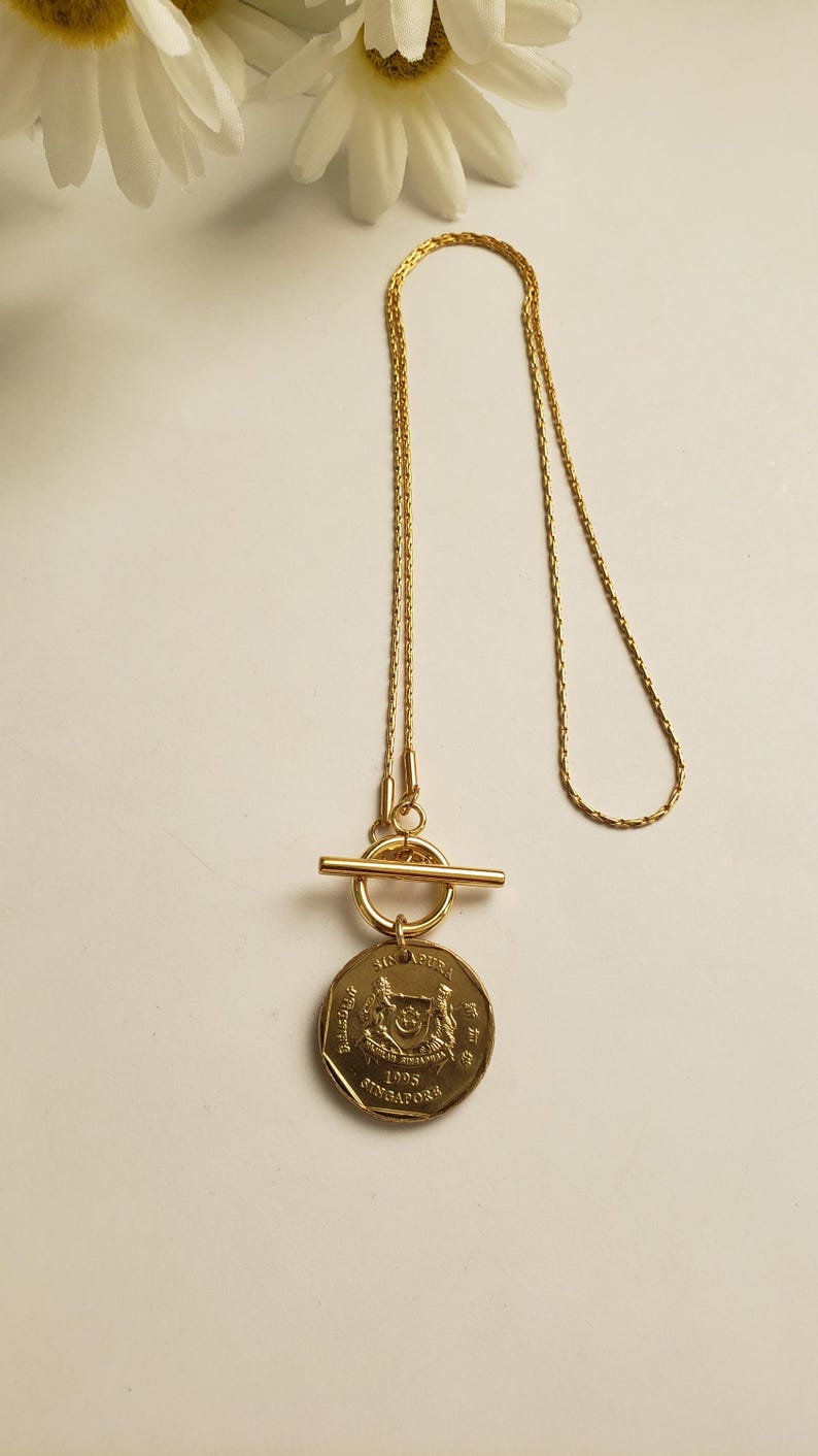May include: A gold necklace with a toggle clasp and a Singapore dollar coin pendant. The coin features the Singapore coat of arms and the year 1995.