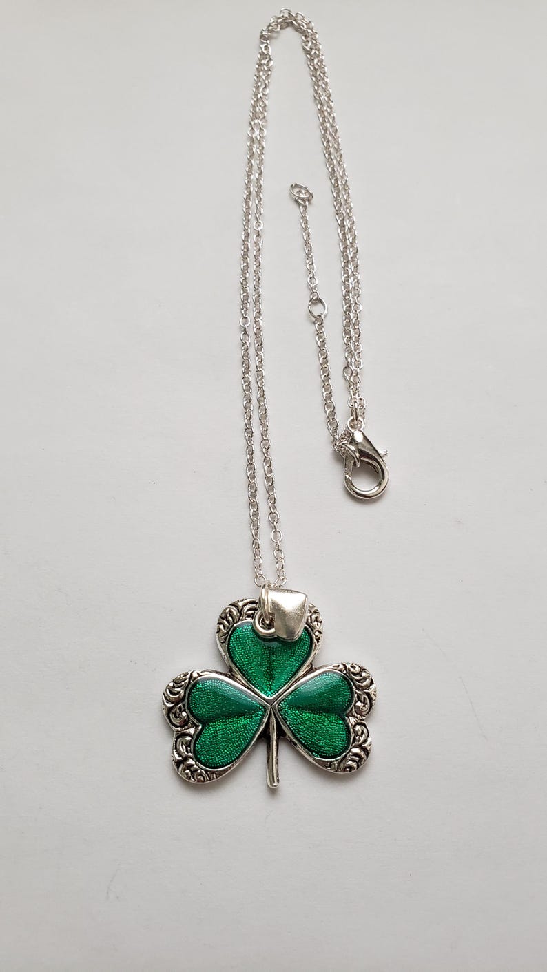May include: A silver chain necklace with a green shamrock pendant. The shamrock is detailed with silver scrollwork and has a green enamel finish.